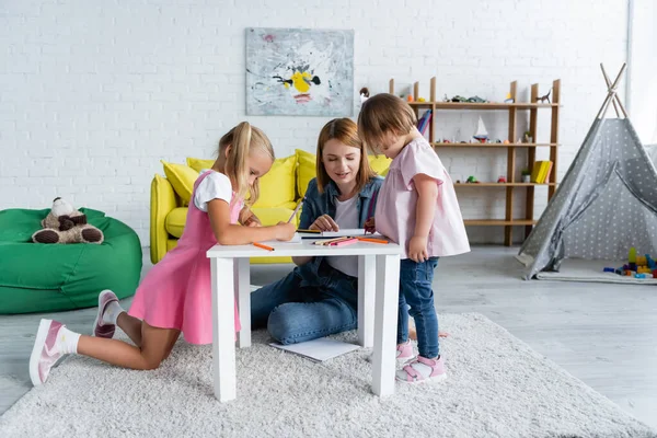 Professeur souriant de maternelle parlant avec tout-petit enfant avec le syndrome du duvet près des papiers et crayons de couleur — Photo de stock