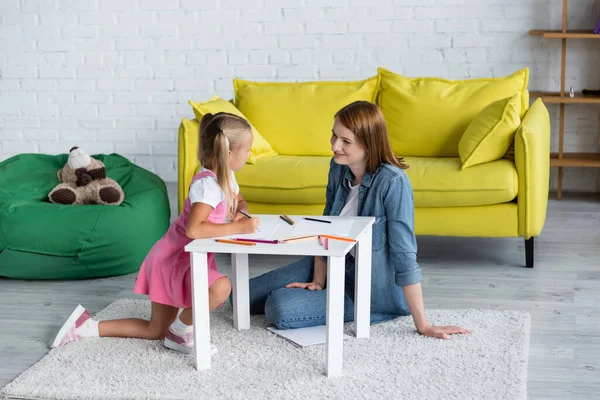 Souriant maternelle enseignant regardant fille près de papiers et crayons de couleur — Photo de stock