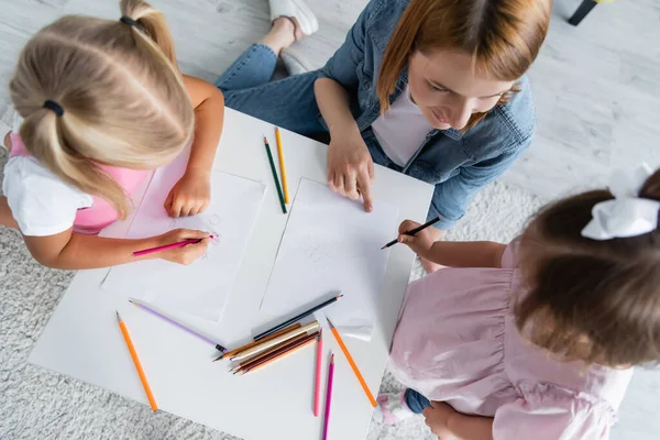 Top view of kindergarten teacher looking at kid with down syndrome near preschooler girl drawing on paper — Stock Photo