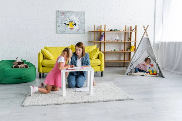 Maestra de jardín de infantes mirando a la chica dibujando mientras niño pequeño con síndrome de Down sentado en tipi - foto de stock