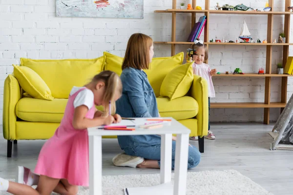 Kindergarten teacher looking at kid with down syndrome playing with toy gun while blurred preschooler girl drawing on paper — Stock Photo