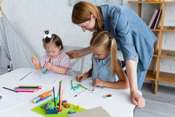 Teacher looking at preschooler girl and disabled child with down syndrome molding plasticine in private kindergarten — Stock Photo