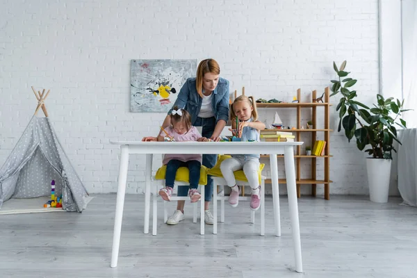 Teacher standing behind preschooler girl and disabled child with down syndrome drawing in private kindergarten — Stock Photo