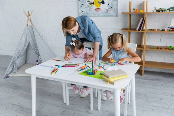 Maestra ayudando a chica discapacitada con síndrome de Down dibujo cerca de preescolar moldeo infantil plastilina en el jardín de infantes privado - foto de stock