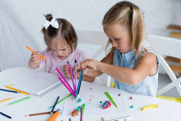 Blonde girl reaching color pencils near disabled toddler kid with down syndrome in private kindergarten — Stock Photo