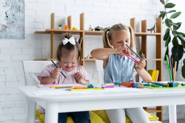 Blonde girl reaching color pencils near disabled toddler child with down syndrome in private kindergarten — Stock Photo