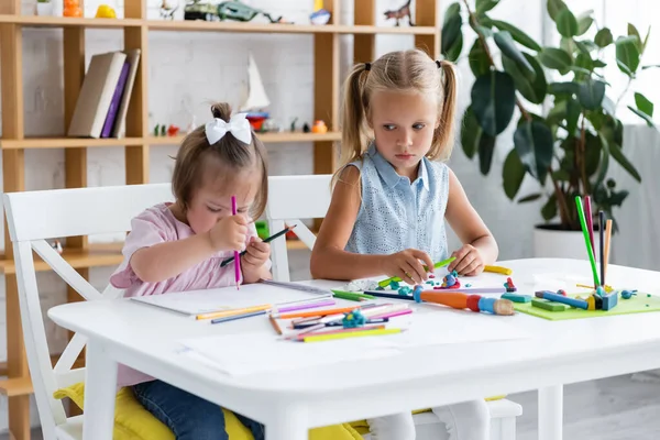 Blonde girl looking at disabled toddler child with down syndrome drawing in private kindergarten — Stock Photo