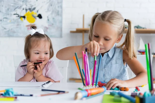 Bambino biondo che raggiunge matite di colore vicino al bambino disabile con sindrome di Down nella scuola materna privata — Foto stock