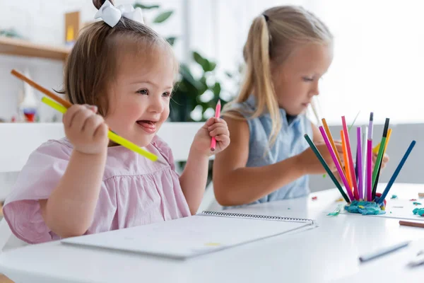 Niño feliz niño con síndrome de Down sosteniendo lápices cerca borrosa chica rubia - foto de stock