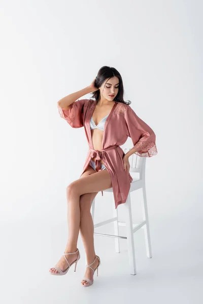Jovem mulher em roupa interior sexy e robe de cetim sentado na cadeira no branco — Fotografia de Stock