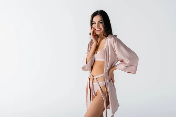 Sorrindo jovem mulher em roupa íntima e robe de seda em pé com a mão no quadril isolado no branco — Fotografia de Stock