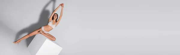 Young woman with closed eyes in white lingerie posing with raised hands on cube on grey, banner — Stock Photo