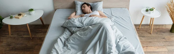 Brunette homme dormant dans le lit sous couverture bleue près des tables de chevet, bannière — Photo de stock