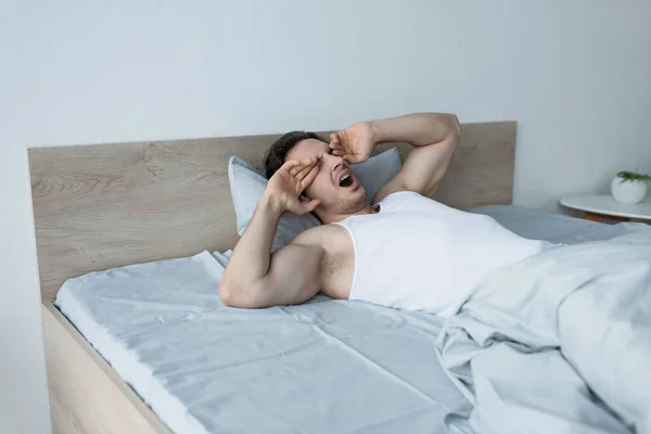 Hombre soñoliento bostezando y frotando los ojos mientras está acostado en la cama por la mañana - foto de stock