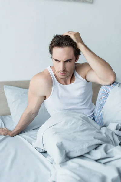 Hombre deprimido sosteniendo la mano cerca de la cabeza mientras está sentado en la cama por la mañana - foto de stock