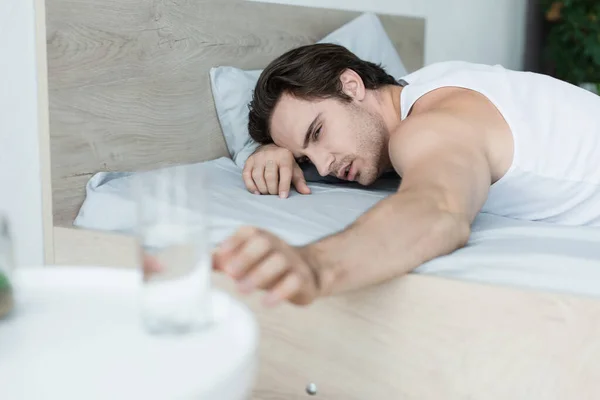 Erschöpfter Mann greift im Bett nach verschwommenem Glas Wasser — Stockfoto