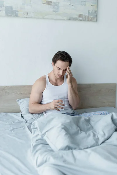 Jovem sentado na cama com copo de água e sofrendo de dor de cabeça — Fotografia de Stock