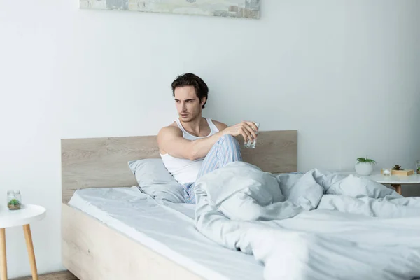 Hombre frustrado sentado en la cama con un vaso de agua y mirando hacia otro lado - foto de stock