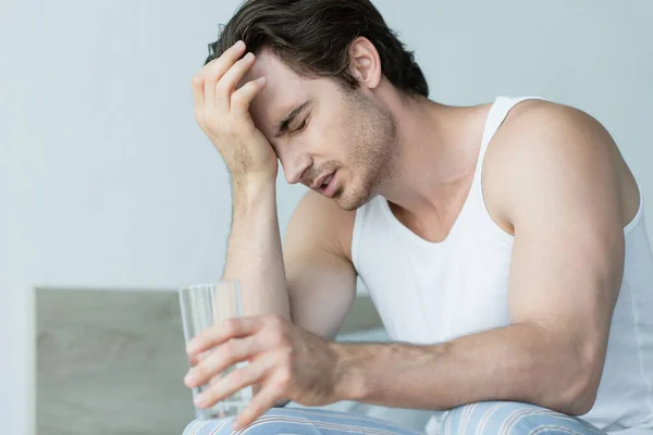 Young man holding glass of water while suffering from headache with closed eyes — Stock Photo