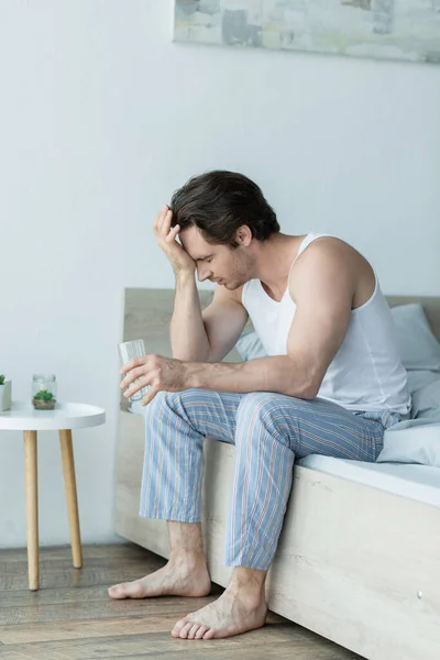 Mann im Schlafanzug leidet unter Kopfschmerzen, während er mit einem Glas Wasser im Bett sitzt — Stockfoto