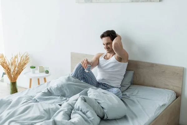 Stirnrunzeln Mann hält Glas Wasser und berührt Kopf, während er im Schlafzimmer an Migräne leidet — Stockfoto