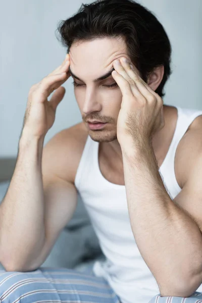 Junger Mann sitzt im Schlafzimmer und leidet unter Kopfschmerzen mit geschlossenen Augen — Stockfoto