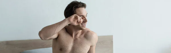 Hombre despierto sin camisa frotando el ojo y sonriendo en el dormitorio, pancarta - foto de stock