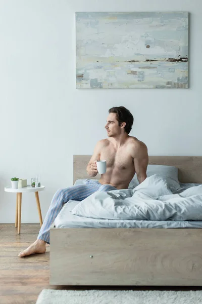 Без сорочки чоловік дивиться геть, сидячи на ліжку з чашкою кави — Stock Photo