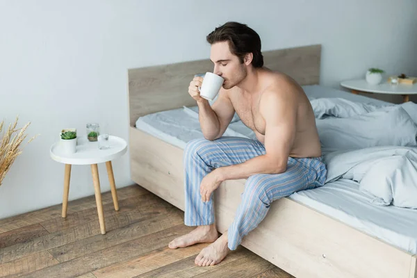 Joven hombre sin camisa en pantalones de pijama rayados bebiendo café de la mañana con los ojos cerrados - foto de stock