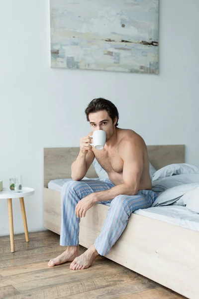 Hemdloser Mann in gestreifter Pyjamahose schaut in die Kamera, während er im Schlafzimmer Kaffee trinkt — Stockfoto