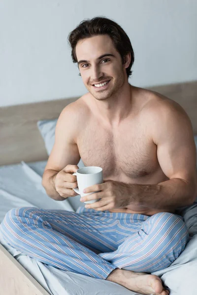 Мужчина без рубашки улыбается в камеру, сидя в постели с чашкой кофе — стоковое фото