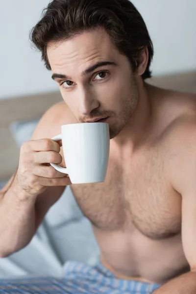 Junger hemdloser Mann trinkt Morgenkaffee und schaut in die Kamera im Schlafzimmer — Stockfoto
