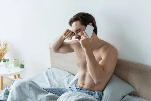 Man rubbing closed eyes while talking on mobile phone in bed — Stock Photo