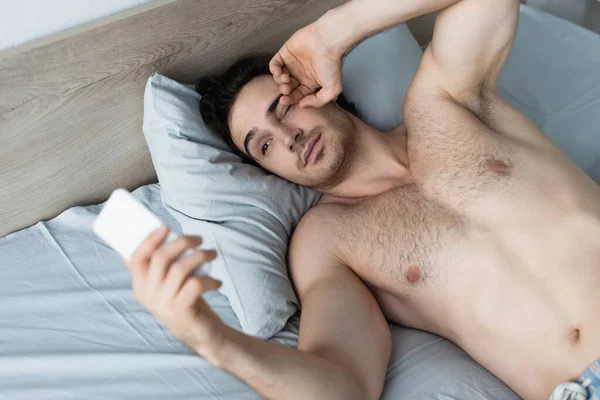 Молодой человек смотрит на размытый смартфон, растирая глаза в постели — стоковое фото