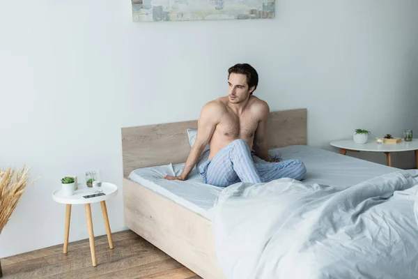 Joven hombre sin camisa sentado en la cama por la mañana y mirando hacia otro lado - foto de stock