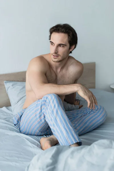 Junger hemdloser Mann in gestreifter Pyjamahose, der im Bett wegschaut — Stockfoto