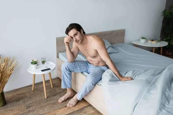 Мужчина без рубашки в синих пижамных штанах чувствует себя нездоровым, сидя на кровати утром — стоковое фото