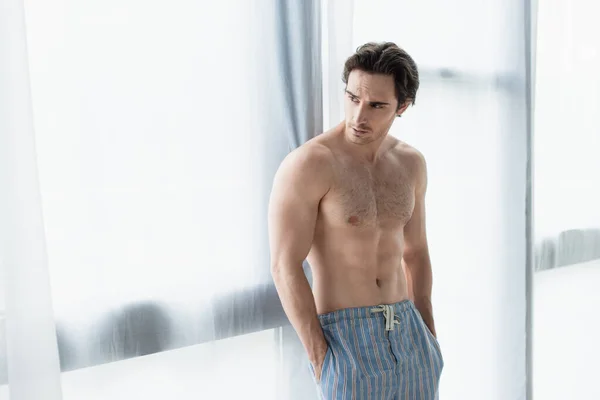 Shirtless man standing near window with hands in pockets of pajama pants — Stock Photo