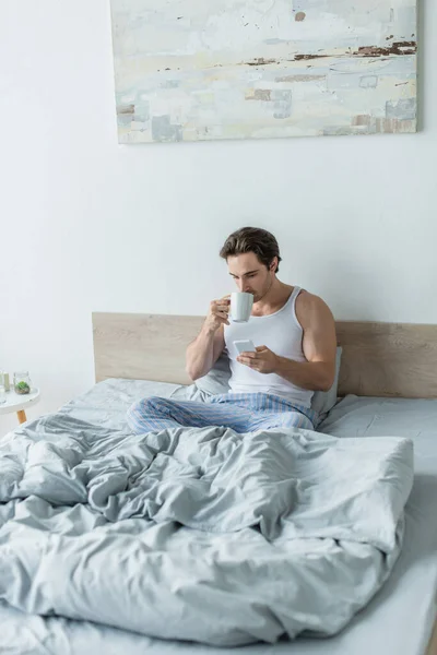 Giovane seduto a letto, che beve caffè e messaggia sul cellulare — Foto stock