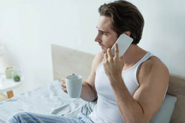 Hombre frunciendo el ceño con taza de café hablando en el teléfono móvil en el dormitorio - foto de stock