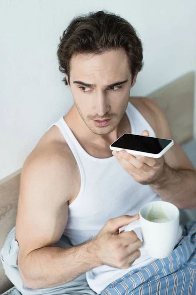 Brunette man holding cup of coffee and sending voice message on smartphone in bedroom — Stock Photo