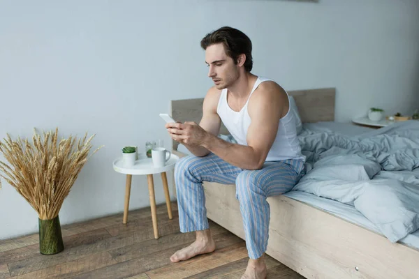 Uomo seduto sul letto vicino al comodino e chatta sul cellulare — Foto stock