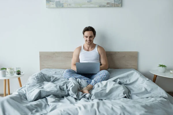 Hombre sonriente sentado en la cama y usando el ordenador portátil - foto de stock