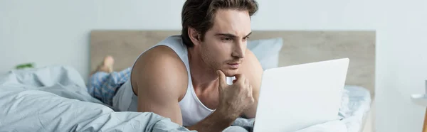Homme réfléchi regardant ordinateur portable tout en étant couché dans le lit, bannière — Photo de stock