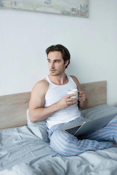 Joven mirando hacia otro lado mientras está sentado en la cama con el ordenador portátil y la taza de café - foto de stock