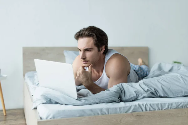Jovem pensando ao usar laptop na cama — Fotografia de Stock
