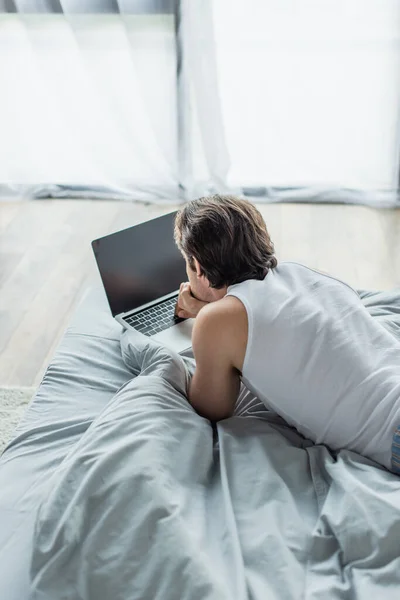 Brunetta uomo sdraiato a letto e utilizzando il computer portatile con schermo bianco — Foto stock