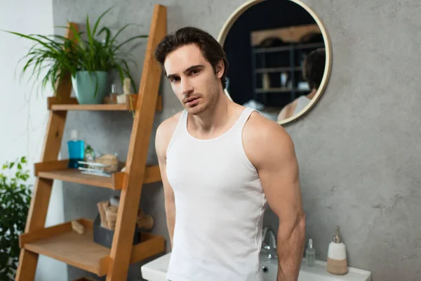Brunette man in white tank top standing near blurred rack in bathroom — Stock Photo