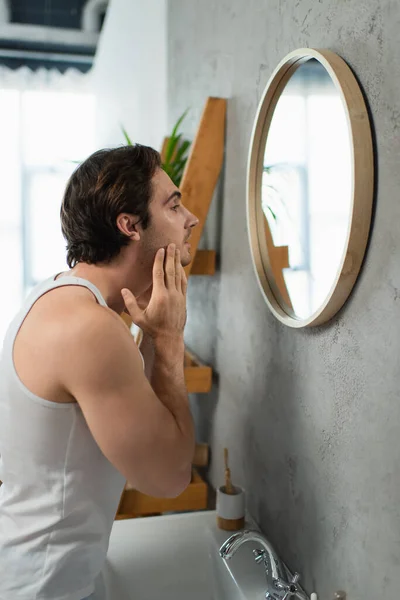 Молодой человек трогает щетину на лице, глядя в зеркало — стоковое фото