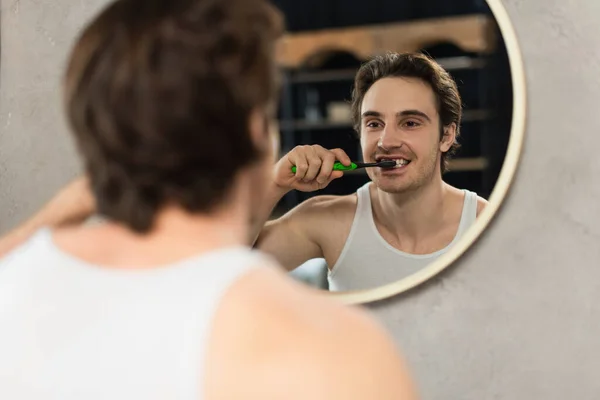 Молодой человек чистит зубы возле зеркала на размытом переднем плане — стоковое фото